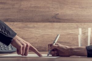 A person signing an agreement with another person pointing to the line where they should sign